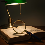 Vintage desk lamp illuminating an open book with a magnifying glass on a dark background